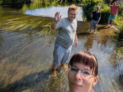 Eine Frau macht ein Selfie im flachen Fluss. Hinter ihr winkt ein Mann fröhlich in die Kamera. Zwei weitere Personen stehen weiter hinten im Wasser. Auf einer Brücke darüber beobachten vier Personen die Szene von oben.