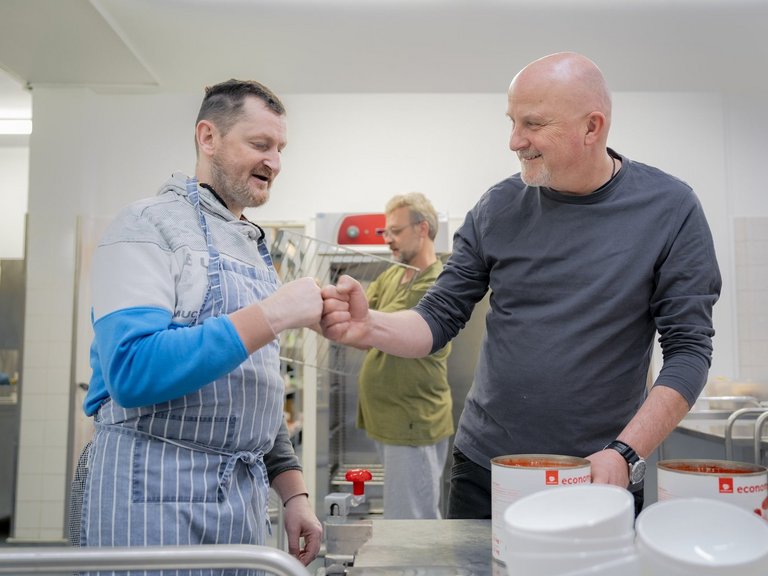 Zwei Männer stehen in einer Großküche vor einigen Dosen Tomatensauce und begrüßen sich per Fistbump.