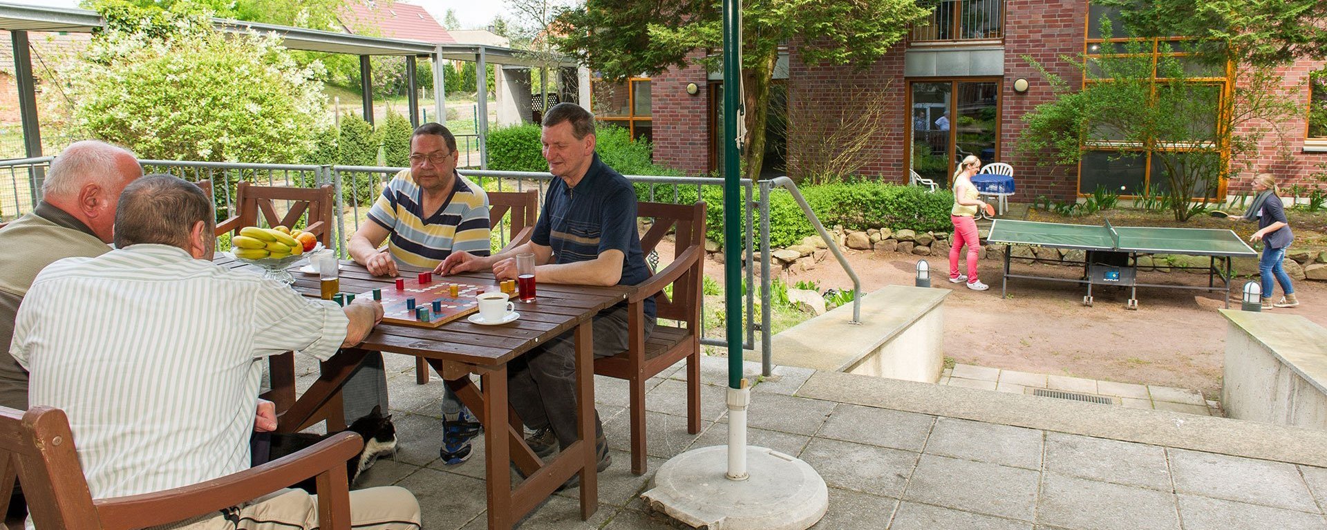 Vier ältere Männer spielen auf der Terasse "Mensch ärgere dich nicht" - im Hintergrund zwei Frauen spielen Tischtennis