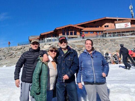 Vier Personen in Winterkleidung stehen nebeneinander im Schnee und lächeln in die Kamera. Im Hintergrund erhebt sich eine große Holzhütte auf einem felsigen Hang unter einem klaren blauen Himmel.