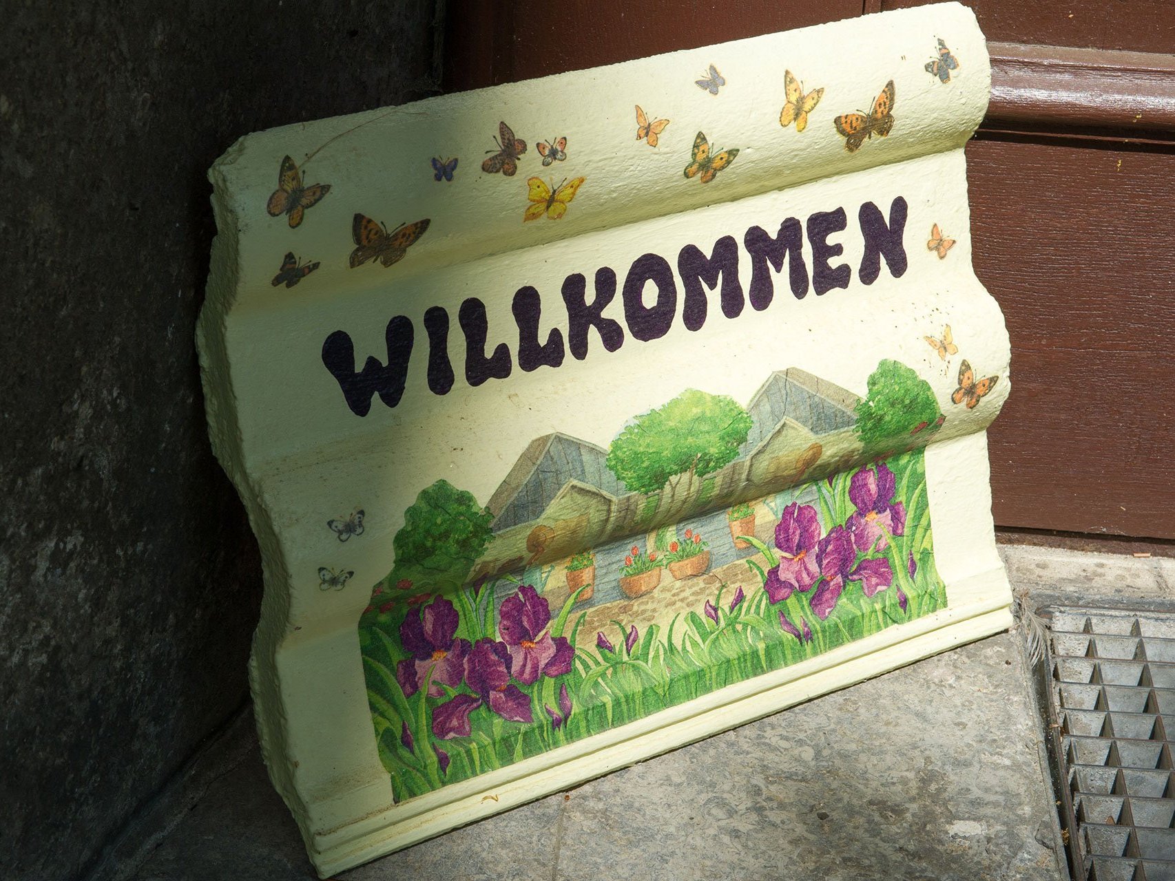 Nahaufnahme: Ein bemalter Dachziegel neben der Eingangstür mit der Aufschrift "WILLKOMMEN", einer gemalten Wiesenlandschaft und bunten Schmetterlingen
