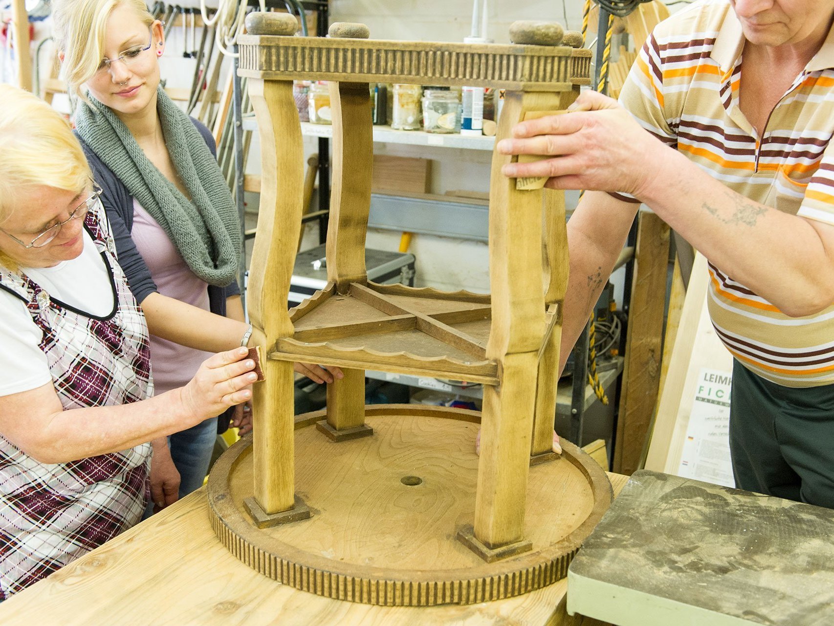 ein alter Holztisch wird von zwei Frauen und einem Mann restauriert - der Mann hält fest, eine Frau "schmirgelt"