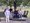 Sieben Personen posieren für ein Gruppenfoto auf einer runden Holzplattform mit Bänken unter einem großen Baum. Im Hintergrund sind eine Wiese und ein Dorf zu sehen.